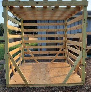 A home made wooden pergola sits in wood chips with grass and a metal building in the background