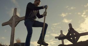 A man holds a shovel in a cemetery surrounded by crosses