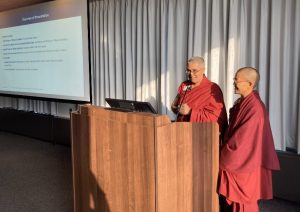 Two Buddhist nuns behind a lectern with a powerpoint slide in the background