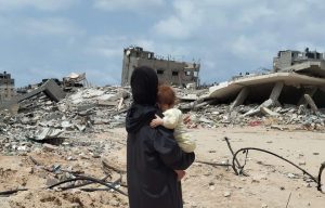 A woman in black holds a baby with rubble and destroyed buildings in the background