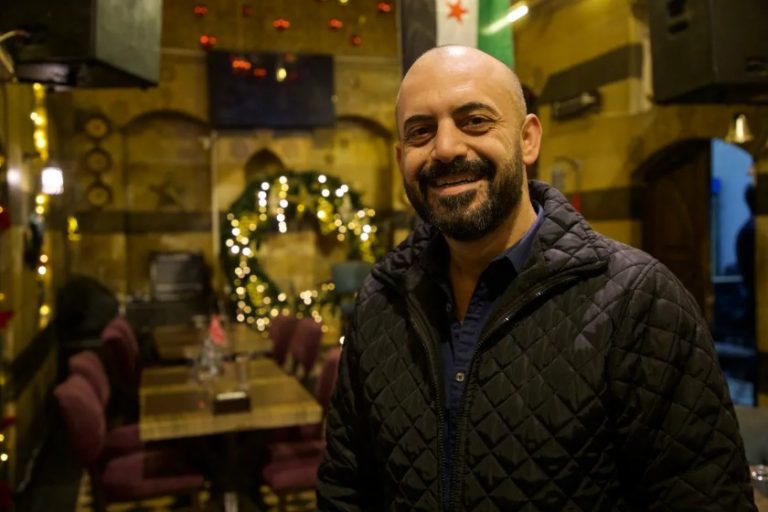 A man smiles with a Christmas wreath and a Syrian flag behind him